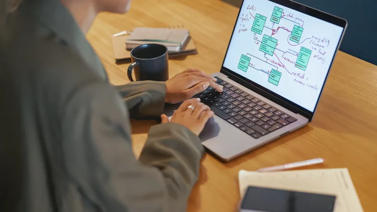 Osoba korzystająca z laptopa z diagramem schematów blokowych lub tabel bazy danych - prawdopodobnie budująca na platformie no-code. Filiżanka kawy, notatnik, długopis i smartfon spoczywają na drewnianym stole. Osoba ma na sobie zieloną kurtkę.