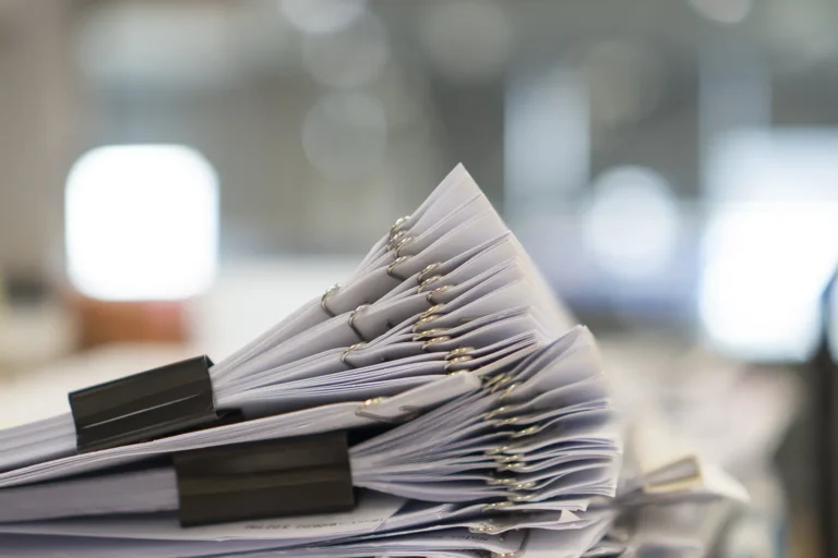 Zbliżenie ułożonych w stos papierowych dokumentów spiętych segregatorem i spinaczami, na rozmytym tle biura - podkreślające potrzebę elektronicznego obiegu dokumentów w nowoczesnych miejscach pracy.